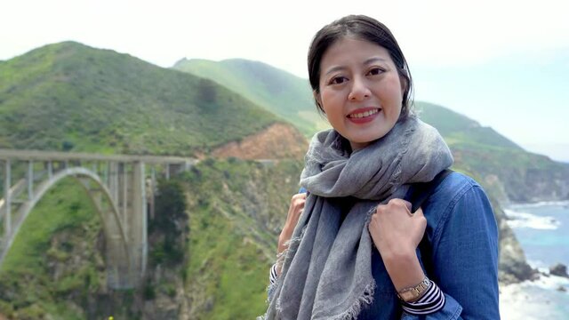 young woman tourist standing in big sur hills and pacific ocean background in california USA. smiling girl traveler face camera with bixby creek bridge on sunny day. female hiker outdoor joyful relax