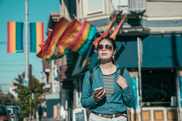 Naklejka na ściany i meble beautiful lady traveler with backpack using cellphone searching way while sightseeing in castro district. woman in sunglasses walking on city street enjoy sunshine. rainbow flags hang on corner store