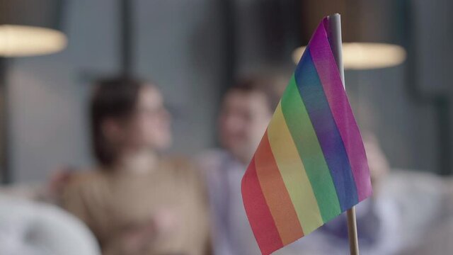 Rainbow lgbt flag at front and blurred gay couple talking at the background. Symbol of same sex love with happy Caucasian boyfriends chatting. Equality, lifestyle.