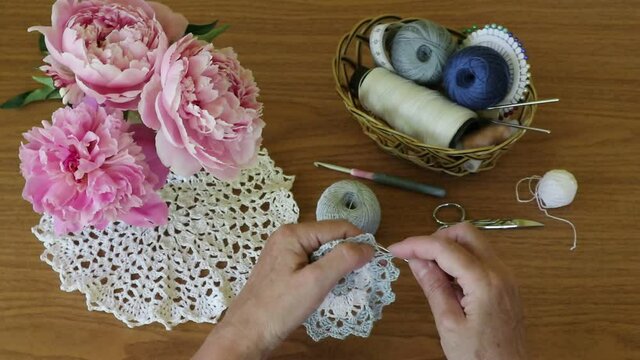 Needlework. Women's hands crochet a lace doily  made of cotton yarn. Vase with peonies on a beautiful napkin, a basket with threads and a set of hooks on a wooden table