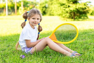 A little girl with ponytails is sitting on the grass with a yellow tennis racket.