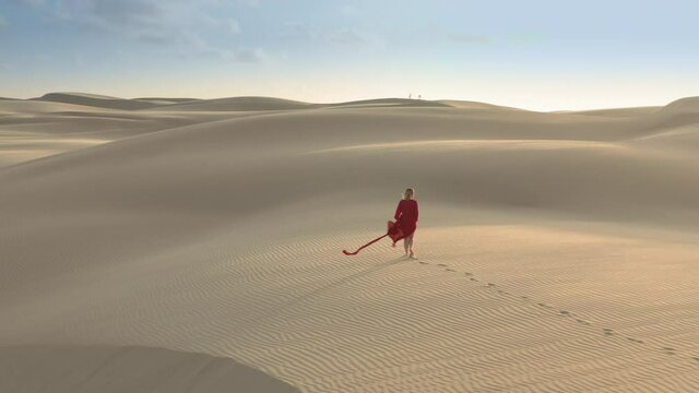 Aerial view of a young model is walking by the natural desert landscape. The fashion concept, long red dress is fluttering on the wind. 4K drone shot of a beautiful woman looking sunset with backlight