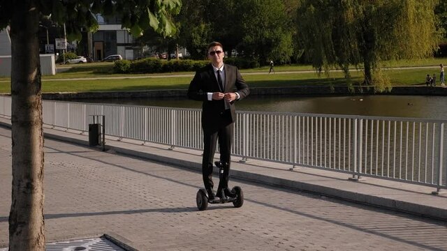 A cheerful man in a business black suit rides a gyro scooter in the Park and buttons up his jacket. Slow motion.