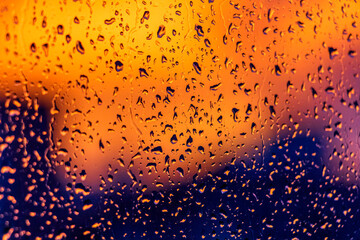 Bright fiery sunset through raindrops on window with bokeh lights. Abstract background. Water drop on the glass against the blurred silhouettes high-rise city.