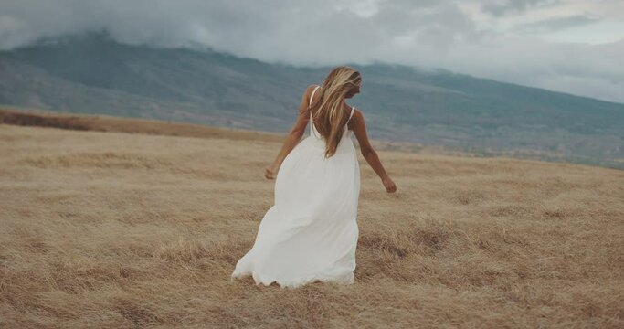 Young beautiful woman dancing in windy field by the mountains in slow motion, bohemian white dress