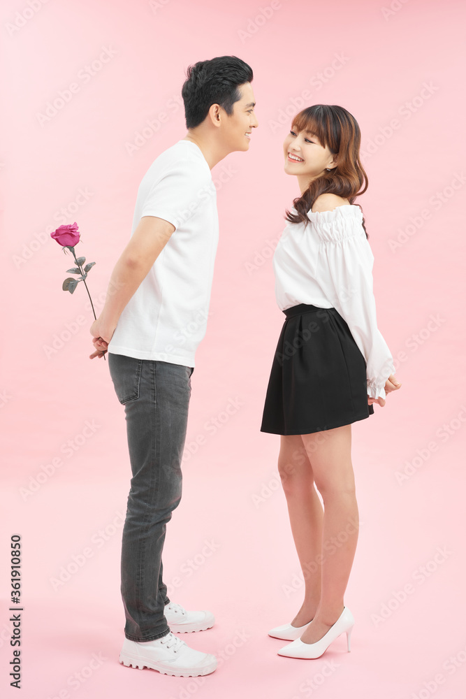 Wall mural First date. Cheerful young loving couple holding a rose and smiling while standing face to face and isolated on pink background - Wall murals