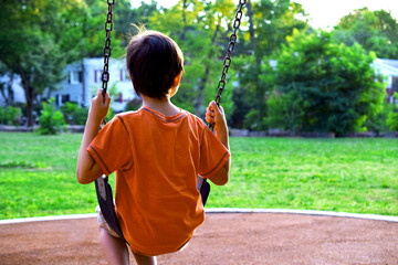 Boy, child sitting on a swing in a playground and looking off into the distance. View from behind, rear.