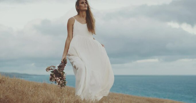 Woman walking through windy field by the ocean in beautiful white dress in slow motion, bohemian chic bouquet