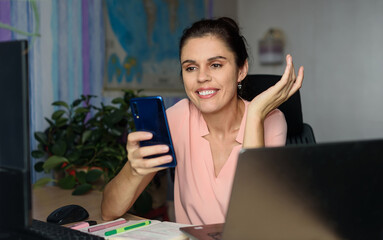 Smiling young woman working home near laptop, shrugging with hand sideway, holds phone, smile...