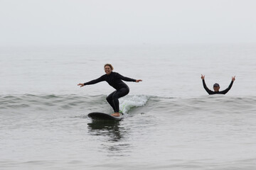 Young multiracial African American lady with amazing smile, freckles & frizzy hair & an Asian Japanese Surf Instructor having a surf lesson together in Chiba, Japan They are wearing black wetsuits.