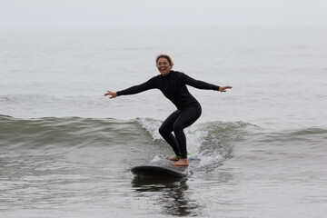 Young multiracial African American lady with amazing smile, freckles & frizzy hair, learning to surf wearing a black wetsuit on a black surfboard. She is Surfing in Chiba Japan near to Ichinomiya.