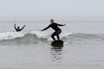 Young multiracial African American lady with amazing smile, freckles & frizzy hair & an Asian Japanese Surf Instructor having a surf lesson together in Chiba, Japan They are wearing black wetsuits.