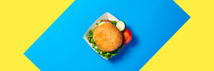 Fresh burger, sliced cucumber, tomato on a yellow-blue background. Flat lay, top view. Banner.