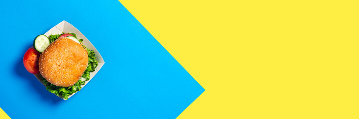 Fresh burger, sliced cucumber, tomato on a yellow-blue background. Flat lay, top view. Banner.