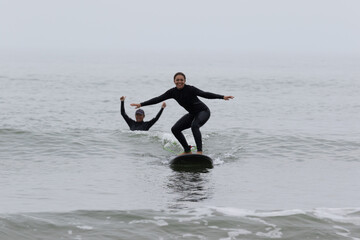 Young multiracial African American lady with amazing smile, freckles & frizzy hair & an Asian Japanese Surf Instructor having a surf lesson together in Chiba, Japan They are wearing black wetsuits.