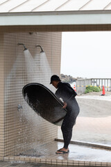 Japanese man in black wetsuit cleaning up his surfboard after surfing on the beach and in the shower. he is wearing a surf cap. In Chiba Japan close to the 2020 surf venue.