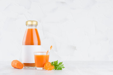 Fresh vegetable orange carrot juice in glass bottle with blank label mock up with glass, straw, green parsley, slices in soft light white interior on wood table.