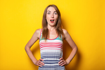 Young woman in a striped sundress surprised in shock on a yellow background.