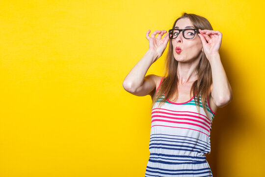 Young girl makes lips bow holding on glasses in a striped dress on a yellow background