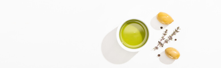 top view of olive oil in bowl near green olives, herb and black pepper on white background, panoramic orientation