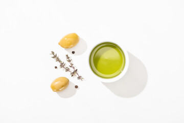 top view of olive oil in bowl near green olives, herb and black pepper on white background