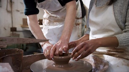 Close up of male teacher helping woman sitting at pottery wheel in ceramics studio to shape clay vase - shot in slow motion