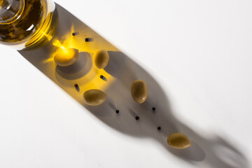 top view of olive oil in bottle near green olives and black pepper in shadow on white background