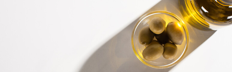 top view of olive oil in bottle near green olives in bowl in shadow on white background, panoramic orientation