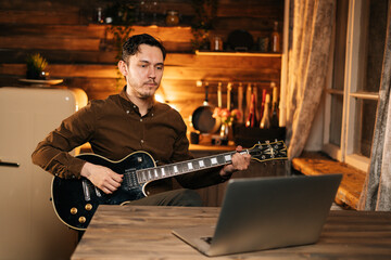 Concentrated young man in casual clothing is watching online tutorial and learning to play guitar using laptop computer at home sitting in dark living room at the desk.