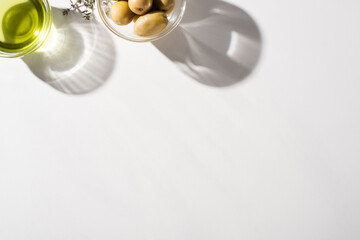 top view of olive oil in bowl near herb, green olives on white background with shadow