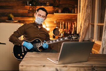 Young man in protective mask and rubber gloves is singing and playing guitar at home, laptop computer on desk, closed eyes. Concept of lifestyle in self-isolation in Coronavirus COVID-19 Pandemic.