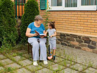 An elderly woman is sitting in the courtyard on a summer day and reading a book with small granddaughter. The concept of a happy old age and International Day of Older Persons and Grandparents' Day