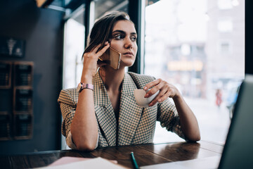 Smart casual manager with coffee cup making business conversation via smartphone application for discussing delivery order, Caucasian female small owner sitting at cafe table with laptop and calling