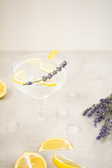 One beautiful glass of lemonade with ice and lemon stands on a light gray concrete surface. Near lie slices of lemon, lavender flowers and melting ice cubes. The glass is decorated with lavender. 