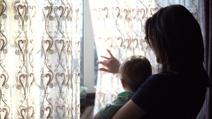 A young mother holds a baby in her arms. A woman with a child looking out the window pushing the curtain.