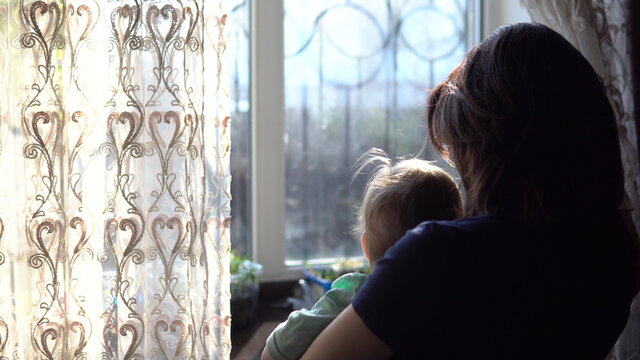 A Young Mother Holds A Baby In Her Arms. A Woman With A Child Looking Out The Window Pushing The Curtain.