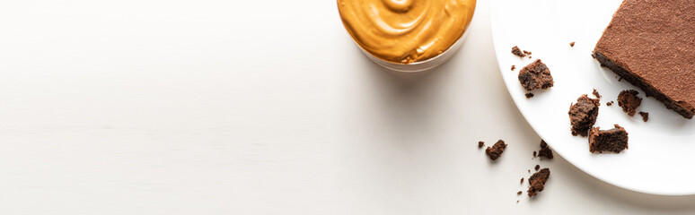 top view of delicious brownie piece on plate near peanut butter on white background, panoramic shot