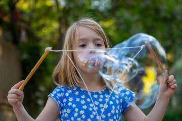 Young girl in a blue and white dress, blowing large bubbles in a backyard setting  © Rachel