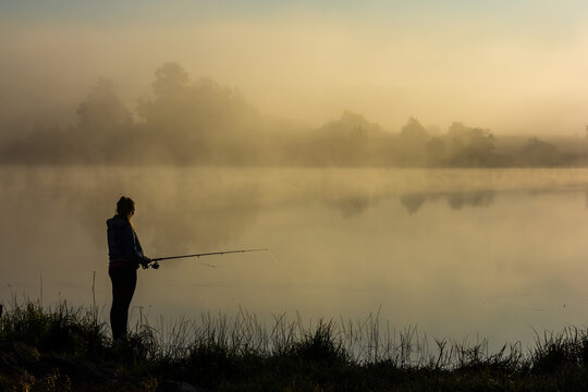 Girl fishermen at dawn. Girl Fishermen in the morning fog on the river bank. Fishermen with fishing rods. Autumn fishing. Morning fog at sunrise. Autumn morning fog.