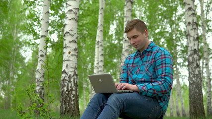 Young man in nature with a laptop in his hands. A man sits on a stump in a birch forest and leads...