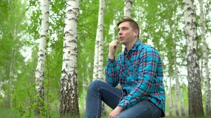 A young man in nature is talking on the phone through headphones. A man sits on a stump in a birch forest with headphones.
