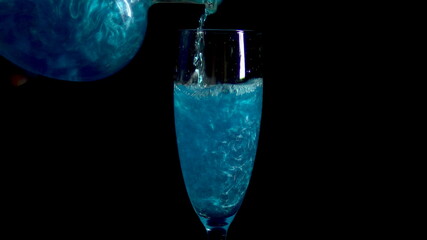 A blue drink is poured into a glass on a black background. The elixir spins and shimmers. Potion of love is isolated on a black background.