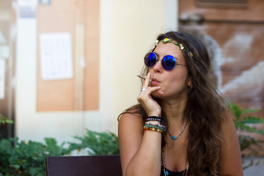 Beautiful Hippy curly brunette girl with round retro blue sunglasses smoking in the street. Woman with cigarette, sunglasses, bracelets, beads, headband looking to the left side. Free gypsy lifestyle