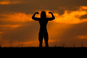 A young bodybuilder woman posing outdoors on the meadow. The clouds are orange and the sun’s rays are visible. silhouette wallpaper nature at sunset. Sport, health and fitness concept