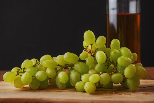 Still life with big cluster of white juicy grapes, bottle of white wine without any label, . Dark black background, Wooden table.