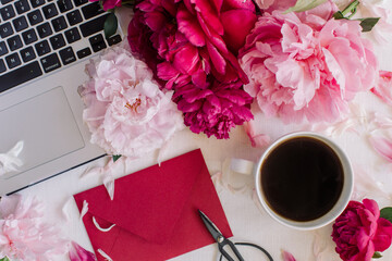 Top view of stylish workspace with laptop, mockup envelope, scissors, cup of coffee and peonies. Wedding invitation concept.