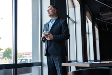 Fototapeta premium Mature businessman drinking water and looking out of a window at the city from an office building.