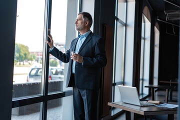 Fototapeta premium Mature businessman drinking water and looking out of a window at the city from an office building.