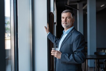 Mature businessman drinking water and looking out of a window at the city from an office building.
