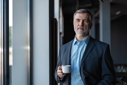 Mature Businessman Drinking A Coffee And Looking Out Of A Window At The City From An Office Building.